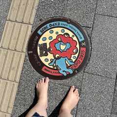 A picture of an Ōsaka Expo 2025-themed manhole cover with the multi-eyed blue and red mascot on it. The tops of my bare feet are visible in the shot as I took the photo pointing directly down.