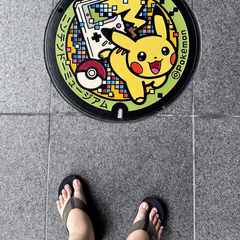 A picture of a Pokémon-themed manhole cover with Pikachu running from a Game Boy and a Pokéball. The tops of my feet, wearing jandals, are visible in the shot as I took the photo pointing directly down.