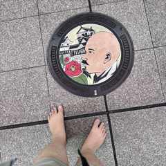 A picture of a Matsuyama-themed manhole cover with a depiction of Masaoka Shiki, a local poet from years ago, next to a Camellia flower in front of Matsuyama Castle. The tops of my bare feet are visible in the shot as I took the photo pointing directly down.