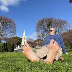 Me sitting on the grass in The Square. In the foreground, I'm sitting with my feet to the camera, leaning back on my palms, looking at something in the distance. In the background, the women's rest can be seen though it is blurry.