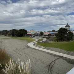 A view of the Wairoa township area from the bridge. In the distance, one can see a few shops in a row, including a large sign for Gemmells (now defunct). To the right, behind a tree, is the Portland lighthouse. In the foreground, a view down to what used to be a large green lawn on the river front, now replaced by a path made of grey silt.