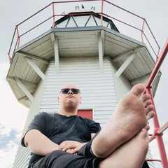 Me sitting on the ground in front of the Portland Island lighthouse in Wairoa. Because I didn't have anybody to hold the camera, the phone camera is on the ground looking upwards at me. The soles of my bare feet are visible in the lower third of the image. I am looking at something out of frame.