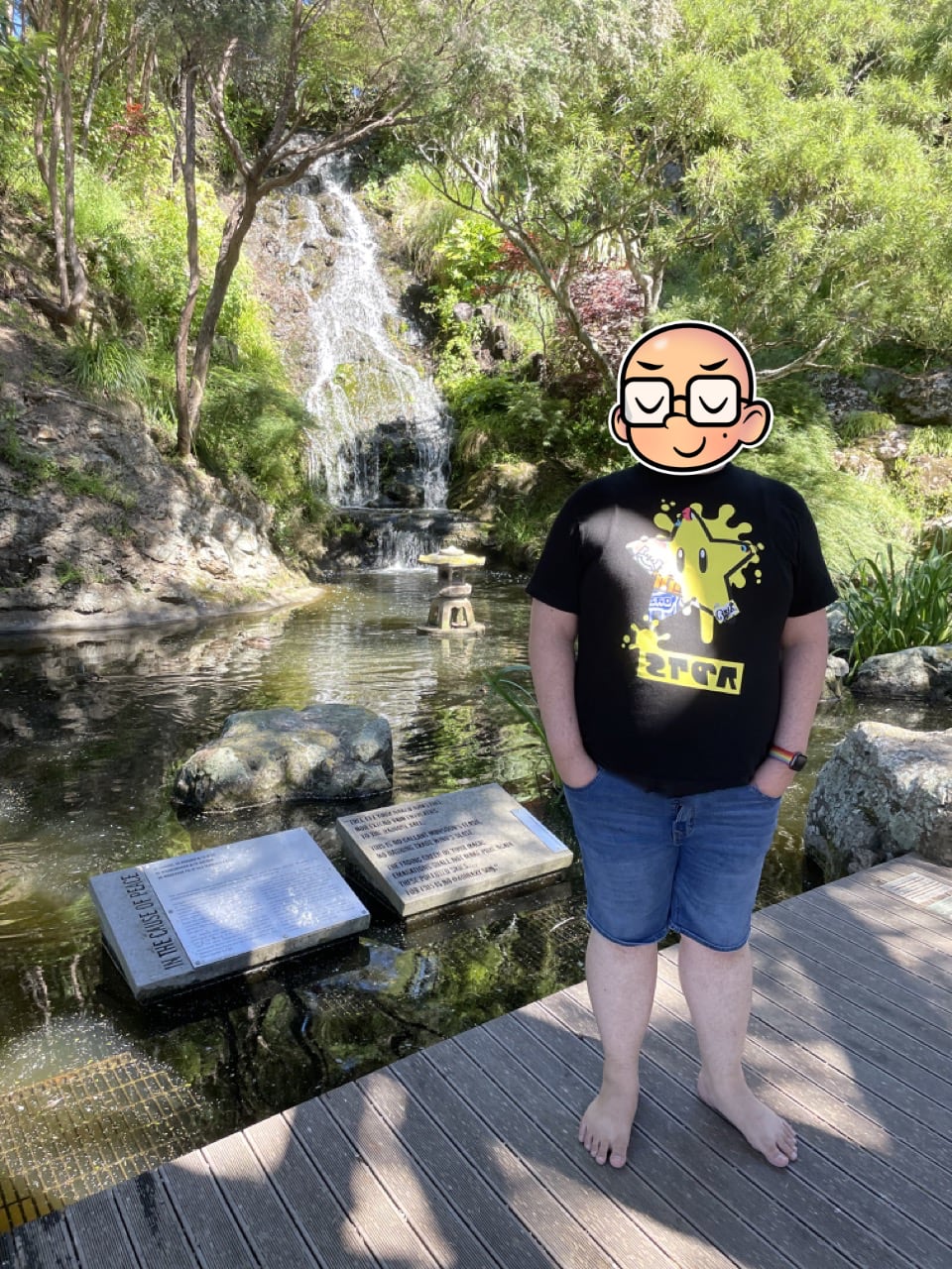 Feets stands in the shade near a pond in the middle of which is a small, Japanese-style lantern which has no flame. A waterfall and some green trees surround the frame of the picture