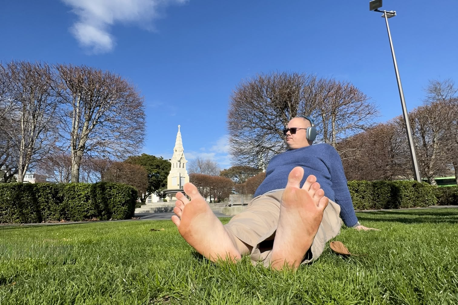 Me sitting on the grass in The Square. In the foreground, I'm sitting with my feet to the camera, leaning back on my palms, looking at something in the distance. In the background, the women's rest can be seen though it is blurry.