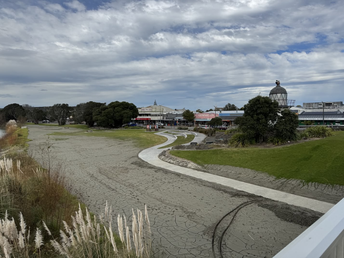 A view of the Wairoa township area from the bridge. In the distance, one can see a few shops in a row, including a large sign for Gemmells (now defunct). To the right, behind a tree, is the Portland lighthouse. In the foreground, a view down to what used to be a large green lawn on the river front, now replaced by a path made of grey silt.