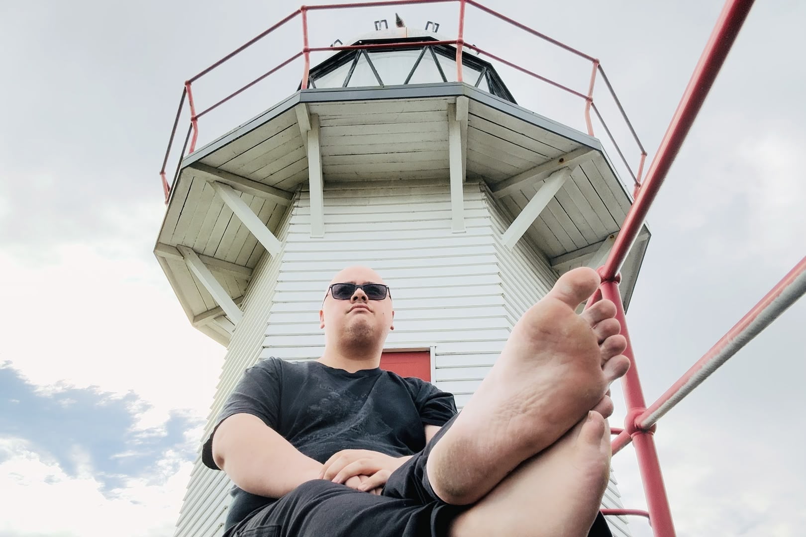 Me sitting on the ground in front of the Portland Island lighthouse in Wairoa. Because I didn't have anybody to hold the camera, the phone camera is on the ground looking upwards at me. The soles of my bare feet are visible in the lower third of the image. I am looking at something out of frame.